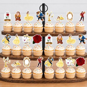 May include: A three-tiered wooden cupcake stand holding twenty-four cupcakes, each topped with a decorative paper cutout featuring characters and icons from Beauty and the Beast, including Belle, the Beast, Mrs. Potts, a red rose, and a mirror.