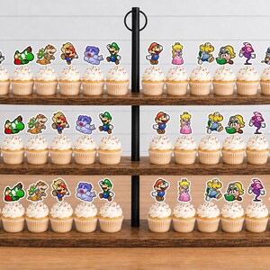 May include: A three-tiered wooden cupcake stand holding numerous cupcakes topped with colorful paper cutouts of various Super Mario characters, including Mario, Luigi, Princess Peach, Yoshi, and other iconic figures from the video game series.