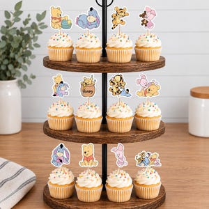 May include: A three-tiered wooden cupcake stand holding twelve cupcakes topped with white frosting and colorful sprinkles. Each cupcake features a paper topper depicting Winnie the Pooh characters including Pooh, Eeyore, Tigger, and Piglet in various poses.