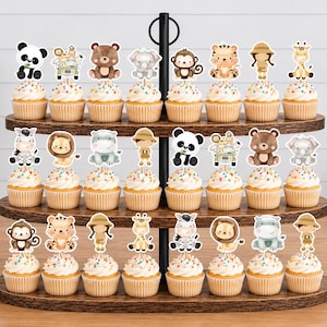 K&ouml;nnte beinhalten: Ein dreist&ouml;ckiger Cupcake-St&auml;nder aus Holz mit vierundzwanzig Cupcakes, die jeweils mit einem dekorativen Papieraufsteller mit Aquarellillustrationen von Dschungeltieren, Safari-Entdeckern und Fahrzeugen auf dem Zuckerguss verziert sind.