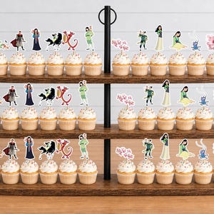 Peut inclure: Un pr&eacute;sentoir en bois &agrave; trois niveaux contenant des rang&eacute;es de cupcakes surmont&eacute;s de d&eacute;coupes en papier d&eacute;coratives repr&eacute;sentant des personnages et des motifs de fleurs de cerisier de l'histoire de Mulan, incluant un guerrier, un dragon et un cheval.