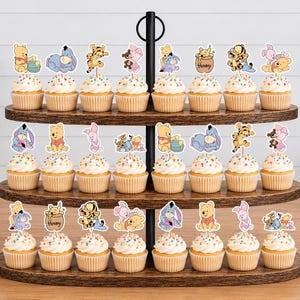 May include: A three-tiered wooden cupcake stand holding twenty-four cupcakes, each topped with a decorative paper cutout featuring Winnie the Pooh, Piglet, Tigger, and Eeyore characters in various poses, with colorful sprinkles on the frosting.
