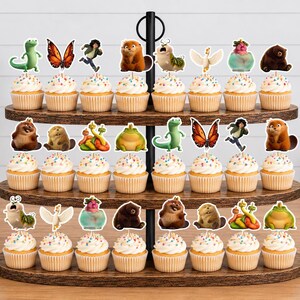 May include: A three-tiered wooden cupcake stand holding twenty-four cupcakes, each topped with a paper cutout featuring various animated characters, including a green lizard, a butterfly, a brown bear, a frog, and a white bird, with sprinkles.