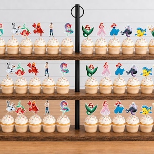 Puede incluir: Un soporte de madera para cupcakes de tres niveles con numerosos cupcakes decorados con recortes de papel coloridos de personajes de La Sirenita, incluyendo a Ariel, Flounder, Sebasti&aacute;n y &Uacute;rsula, dispuestos en filas sobre cada estante marr&oacute;n.