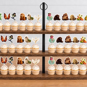 May include: A three-tiered wooden cupcake stand holding eighteen cupcakes, each topped with a decorative paper cutout featuring various colorful cartoon characters, including a green lizard, a butterfly, a person, a beaver, and a frog.