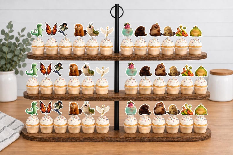 May include: A three-tiered wooden cupcake stand holding eighteen cupcakes, each topped with a decorative paper cutout featuring various colorful cartoon characters, including a green lizard, a butterfly, a person, a beaver, and a frog.