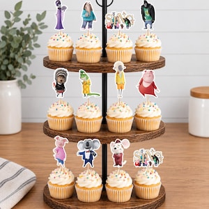 May include: A three-tiered wooden cupcake stand holding twelve cupcakes topped with white frosting and colorful sprinkles. Each cupcake features a paper topper depicting various animated characters from the movie Sing, including the Sing logo text.