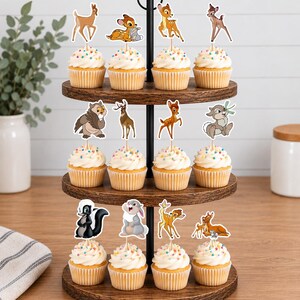 May include: A three-tiered wooden cupcake stand holding twelve cupcakes, each topped with a decorative paper cutout featuring various cartoon forest animals including deer, a rabbit, a skunk, and an owl, all with white frosting and colorful sprinkles.