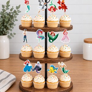 Puede incluir: Un soporte de madera para cupcakes de tres niveles con doce cupcakes decorados con chispas de colores y recortes de papel con personajes de La Sirenita, incluyendo a Ariel, Sebasti&aacute;n, Flounder, el Rey Trit&oacute;n y &Uacute;rsula.