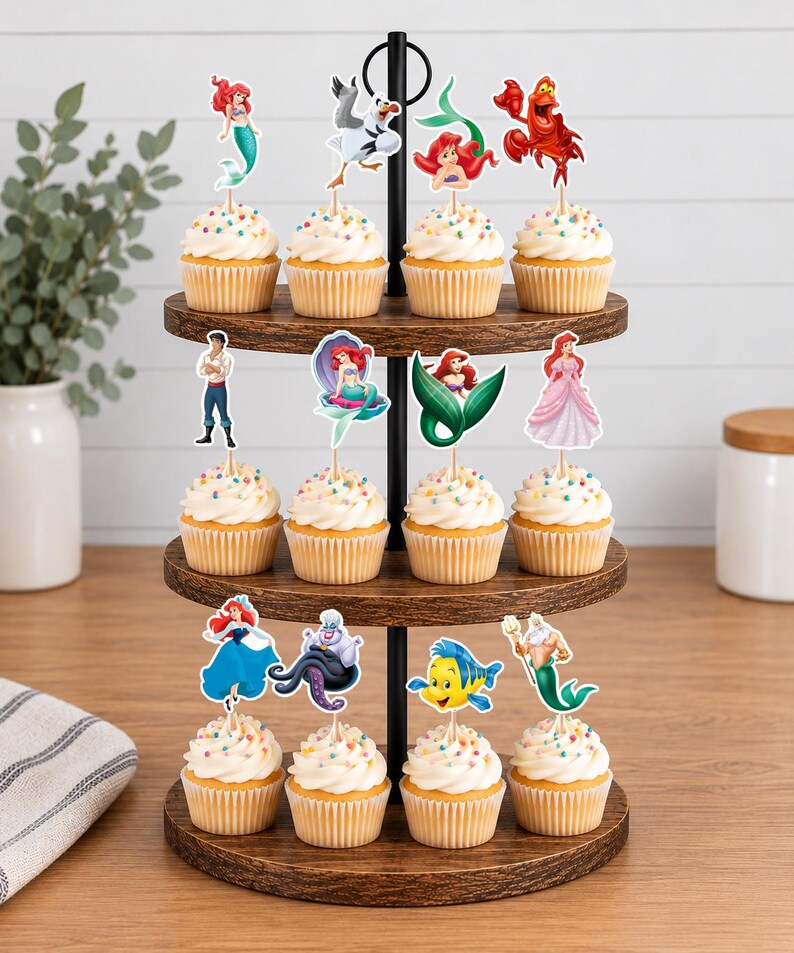 Puede incluir: Un soporte de madera para cupcakes de tres niveles con doce cupcakes decorados con chispas de colores y recortes de papel con personajes de La Sirenita, incluyendo a Ariel, Sebasti&aacute;n, Flounder, el Rey Trit&oacute;n y &Uacute;rsula.