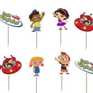 May include: A collection of Little Einsteins cupcake toppers featuring cartoon characters and the show's logo. The toppers include images of the characters Leo, June, Quincy, Annie, and the red rocket ship. Each topper is attached to a wooden stick.