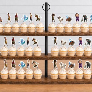 K&ouml;nnte beinhalten: Ein dreist&ouml;ckiger Cupcake-St&auml;nder aus Holz mit Cupcakes, die mit Papieraufstellern von Zeichentrickfiguren verziert sind. Die Figuren zeigen ein kleines Wesen, eine Person in Gelb, eine Person mit rotem Umhang und eine blaue Kreatur.