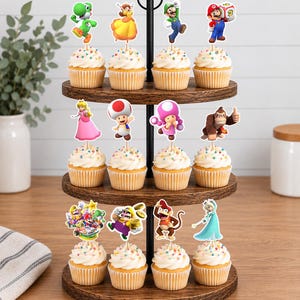 Pu&ograve; includere: Un'alzata per cupcake in legno a tre livelli che sostiene dodici cupcake guarniti con glassa bianca e zuccherini colorati. Ogni cupcake presenta un topper decorativo in carta che raffigura vari personaggi Nintendo tra cui Mario, Luigi, Peach, Toad e Donkey Kong.