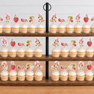 Op de afbeelding: Een houten cupcake-standaard met drie niveaus met talloze cupcakes, bekroond met decoratieve papieren prikkers met illustraties van een meisje met een rode hoed, aardbeien en roze katten. De standaard heeft een zwarte metalen middenstang.