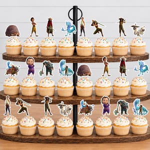 K&ouml;nnte beinhalten: Ein dreist&ouml;ckiger Cupcake-St&auml;nder aus Holz mit vierundzwanzig Cupcakes, die jeweils mit einem bunten Papieraufsteller versehen sind, der verschiedene animierte Figuren aus dem Film Raya und der letzte Drache zeigt, darunter Raya, Sisu und Tuk Tuk.
