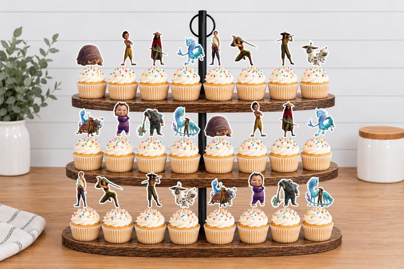 K&ouml;nnte beinhalten: Ein dreist&ouml;ckiger Cupcake-St&auml;nder aus Holz mit vierundzwanzig Cupcakes, die jeweils mit einem bunten Papieraufsteller versehen sind, der verschiedene animierte Figuren aus dem Film Raya und der letzte Drache zeigt, darunter Raya, Sisu und Tuk Tuk.