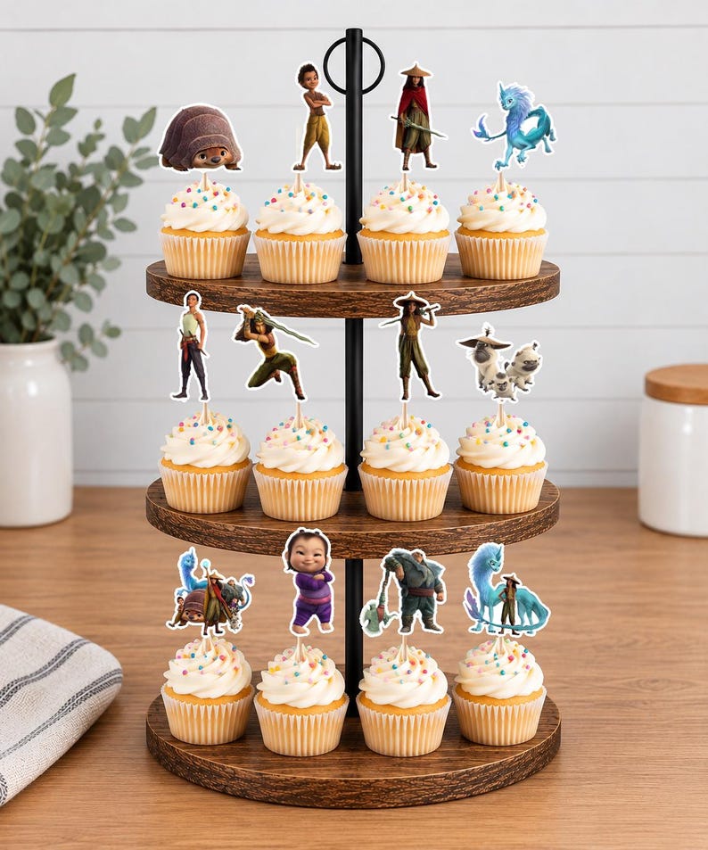 K&ouml;nnte beinhalten: Ein dreist&ouml;ckiger Cupcake-St&auml;nder aus Holz mit zw&ouml;lf Cupcakes, die mit wei&szlig;em Zuckerguss und bunten Streuseln verziert sind. Jeder Cupcake hat einen dekorativen Papieraufsatz, der verschiedene animierte Figuren aus dem Film Raya und der letzte Drache zeigt.