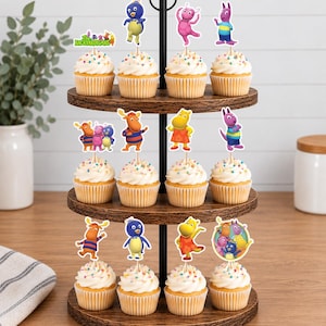 May include: A three-tiered wooden cupcake stand holding twelve cupcakes topped with white frosting and colorful sprinkles. Each cupcake features a paper topper depicting characters from The Backyardigans, including Pablo, Tyrone, Uniqua, Tasha, and Austin.