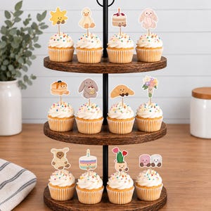 Puede incluir: Un soporte de madera para cupcakes de tres niveles con doce cupcakes cubiertos de glaseado blanco, chispas de colores y varios adornos de papel decorativos con ilustraciones de animales, comida, flores y pasteles.