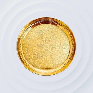 May include: A circular, gold-coloured serving tray featuring an elaborate floral pattern engraved on its surface. The tray has a raised edge with a detailed, ridged design. The overall aesthetic is ornate and decorative, suitable for serving or display.