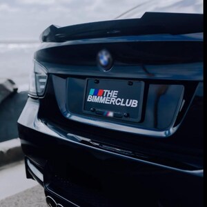 May include: A black BMW car's rear with a spoiler and dual exhaust pipes. The license plate reads "THE BIMMERCLUB" with a colorful logo. The car is parked near the coast, with the ocean visible in the background.