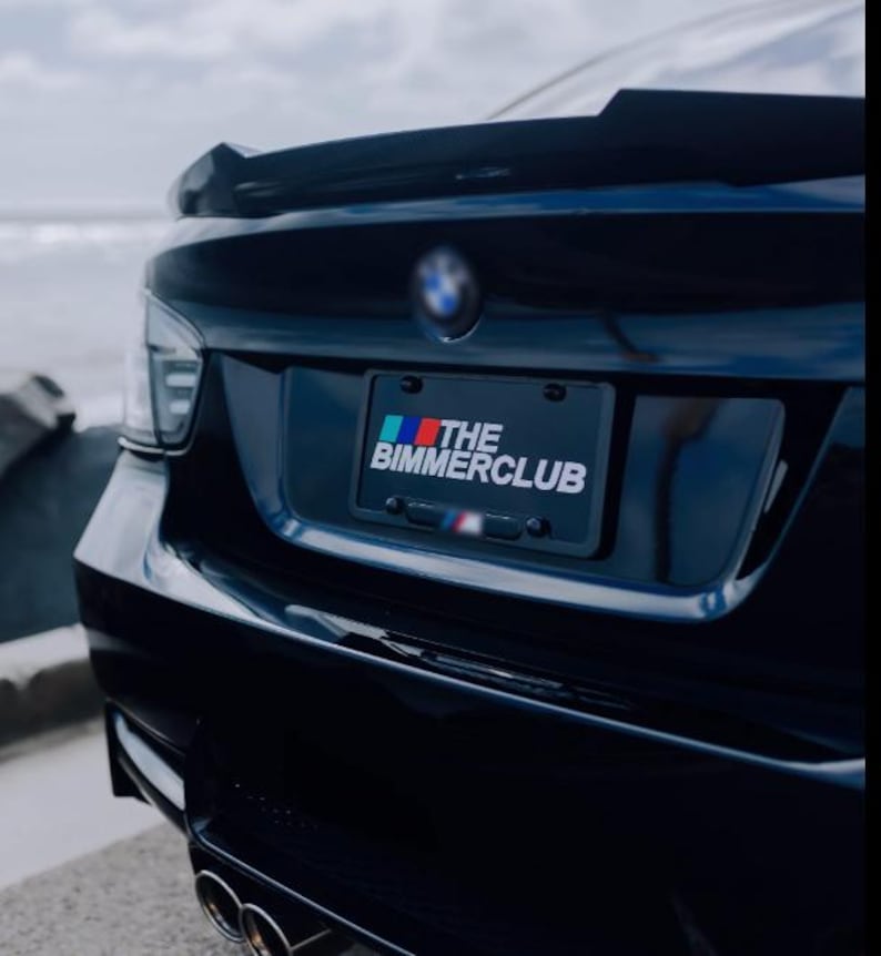 May include: A black BMW car's rear with a spoiler and dual exhaust pipes. The license plate reads "THE BIMMERCLUB" with a colorful logo. The car is parked near the coast, with the ocean visible in the background.