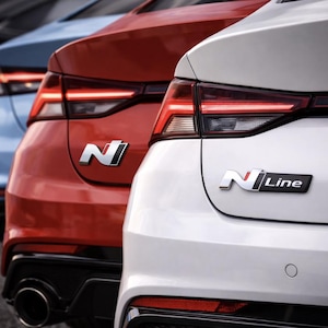 May include: Close-up of the rear of three cars in red, white, and blue. Each car has a distinct taillight design and a chrome emblem. The white car features an "N Line" badge.
