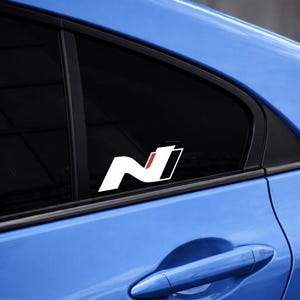 May include: White and red "N" logo sticker on a blue car window. The sticker features a stylized "N" design with a red accent and a black outline. The car window is tinted black.