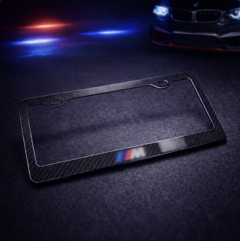 May include: Black carbon fiber license plate frame with the BMW M logo in blue, red, and white. The frame has a textured appearance and a rectangular shape. The background is dark with a car in the background.