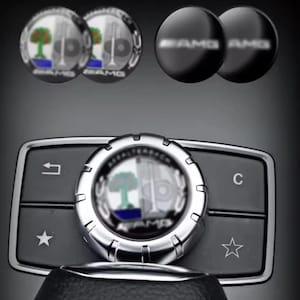 May include: A set of automotive interior accessories including two circular emblems with a tree and valve graphic, two black circular caps with AMG text, and a central control dial featuring the Affalterbach crest, back arrow, and star icons.