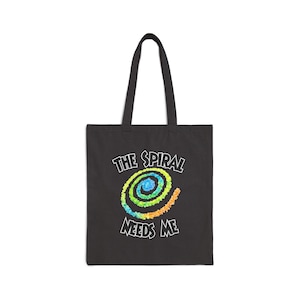 May include: A black tote bag featuring a colorful spiral graphic in shades of blue, green, yellow, and orange. The text "THE SPIRAL" is printed in white above the spiral, and the text "NEEDS ME" is printed in white below the spiral.