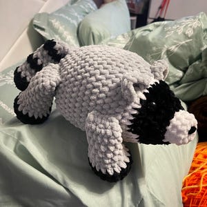 May include: A plush raccoon toy made of soft, light gray yarn with a textured, chunky knit appearance. It features a black mask around the eyes, a white muzzle with a small black nose, black paws, and a striped tail with alternating black and gray bands.
