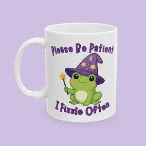 May include: White ceramic mug featuring a cartoon frog wearing a purple wizard hat with stars and a crescent moon. The frog holds a star wand. Text on the mug reads "Please Be Patient I Fizzle Often" in purple.