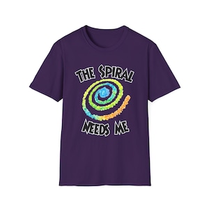 May include: A purple t-shirt with the text "THE SPIRAL NEEDS ME" in white, above a colorful spiral design. The spiral features blue, green, yellow, and orange hues. The shirt is a short-sleeved crew neck.