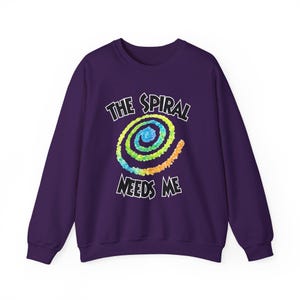 May include: A purple sweatshirt with the text "THE SPIRAL NEEDS ME" in white. The design features a colorful spiral in shades of blue, green, yellow, and orange, centered on the front of the sweatshirt. The sweatshirt has long sleeves.