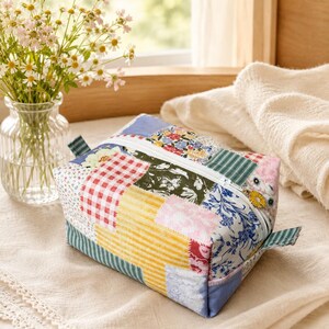 Peut inclure: Une trousse rectangulaire à fermeture éclair avec un motif patchwork composé de divers tissus, dont des carreaux vichy rouges et blancs, des rayures jaunes, des imprimés floraux et des rayures vertes. La tirette est une petite marguerite.