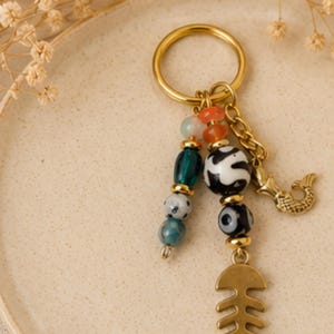May include: A gold-toned metal keychain featuring a dangling fish skeleton charm and a mermaid tail charm. The keychain is adorned with a variety of colorful beads, including teal, blue, orange, and black and white patterned glass beads with gold spacers.