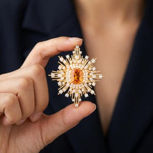 May include: A gold-toned starburst brooch featuring a central rectangular orange gemstone surrounded by numerous small, clear, marquise and round-cut crystals arranged in a radiating pattern that extends outward from the center stone.