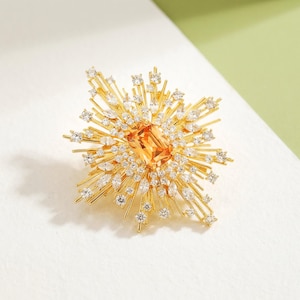 May include: A gold-toned brooch with a starburst design. The center features a rectangular orange gemstone, surrounded by smaller clear crystals and radiating gold bars. The brooch is set against a white and green background.