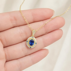 Pode incluir: Um colar dourado com um pingente oval de pedra azul. O pingente é cercado por pedras brancas e azuis menores. O colar é delicado e parece ser de ouro. O pingente tem aproximadamente 2 cm de comprimento.