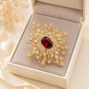 May include: A gold brooch featuring a large, rectangular, deep red gemstone at the center, surrounded by a sunburst arrangement of numerous small, clear, sparkling white stones set on thin, radiating gold-toned metal spikes and prongs.