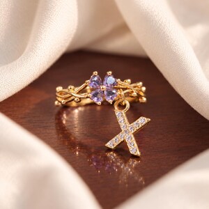 May include: A gold-toned ring featuring a band with a vine-like design, four purple heart-shaped stones arranged in a clover pattern, and a dangling charm in the shape of the letter X, which is encrusted with small, clear, sparkling stones.