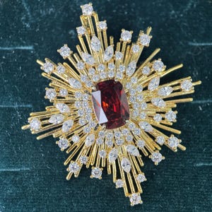 Puede incluir: Un broche dorado en forma de estrella con una gran piedra central rectangular de color rojo intenso, rodeada por múltiples cristales transparentes redondos y de talla marquesa dispuestos en un patrón radiante desde el centro.