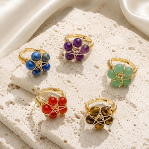 May include: Five gold-toned wire-wrapped rings, each featuring a cluster of gemstone beads in various colors: blue, purple, green, red, and brown. The rings are displayed on a textured, off-white surface.