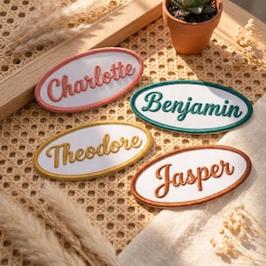 May include: Four embroidered name patches in oval shapes with the names "Charlotte", "Benjamin", "Theodore", and "Jasper". Each patch has a white centre with a coloured border and matching embroidered text. A small potted succulent plant is in the background.