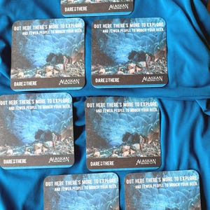 May include: Square coasters with a blue and gray landscape design, featuring text that reads "OUT HERE THERE'S MORE TO EXPLORE AND FEWER PEOPLE TO MOOCH YOUR BEER." The coasters also display the "ALASKAN" logo.