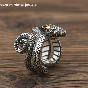 May include: A silver-colored snake ring with a detailed scale pattern. The snake's head is accented with gold-colored metal and yellow-toned eyes. The ring is displayed on a wooden surface. The text "nova minimal jewels" is in the upper left corner.