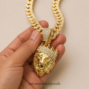 May include: A gold-colored lion pendant necklace with a crown, set with small, sparkling stones. The chain is a thick, gold-colored curb link style. The necklace is held in a hand against a neutral background. The text "novaminimaljewels" is at the bottom.