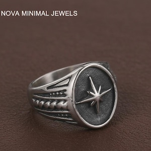 May include: A silver ring featuring a compass rose design. The ring has a circular face with a starburst pattern and the text "NOVA MINIMAL JEWELS" at the top. The band has decorative detailing.