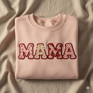 Op de afbeelding: Een lichtroze sweatshirt met het woord "MAMA" geborduurd op de voorkant. De letters zijn rood omlijnd en gevuld met verschillende patronen, waaronder harten, bloemmotieven en stippen. Het sweatshirt is netjes opgevouwen.